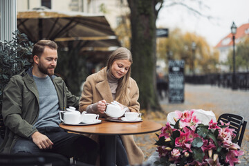 A couple enjoying tea at a cozy outdoor café. The man is wearing a green jacket and the woman is in a beige coat, pouring tea into cups. The setting features autumn leaves and a floral arrangement on