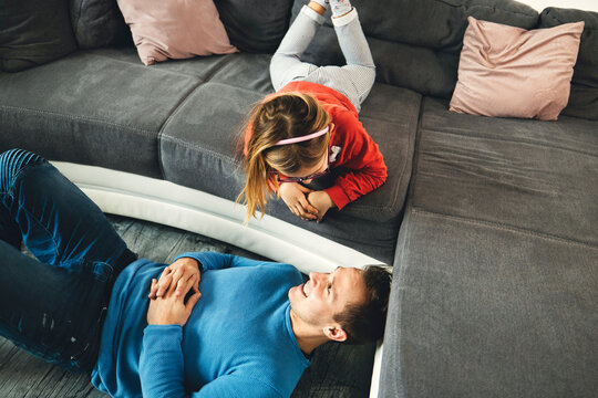 A father and two daughters are joyfully jumping on a couch in a bright living room. The father is mid-air, playfully pointing, while the girls are laughing and leaping. The scene captures a moment of