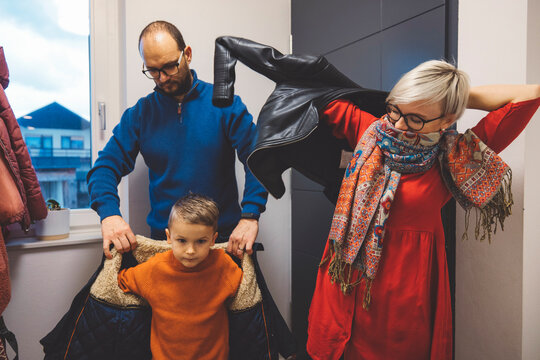 A family preparing to go out, with a father helping a young boy put on a coat while a mother is taking off her jacket. The scene is cozy and homely, showcasing family bonding.