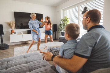 Fototapeta premium A lively family scene in a modern living room. A young girl and an mother are playfully dancing and pointing, while a man sits on the couch, smiling and watching. The room is bright and stylish, with