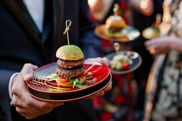 Man waiter is holding a plate with a hamburger on it. The burger is on a bun and has a green topping. The man is wearing a suit and tie