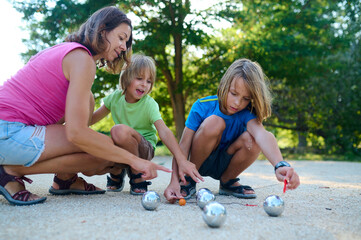 A mother and her two sons measure the distance between petanque balls and the target ball. A fun moment of focus and teamwork during an outdoor family game in a sunny park