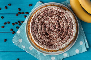 Top view of banoffe pie filled with banana and dulce de leche and sprinkled with grated chocolate. Banana cake on white plate with glass of coffee on turquoise summer background. Flat lay