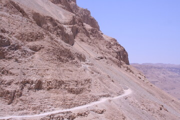 A winding path leads through arid landscapes beneath the sunny sky at Masada, Israel, during midday, showcasing ancient rock formations