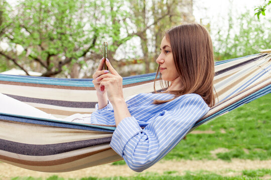 young cheerful girl lies on comfortable hammock in garden in summer and uses smartphone online, woman relaxes in park outdoors and looks at mobile phone and smiles - Powered by Adobe