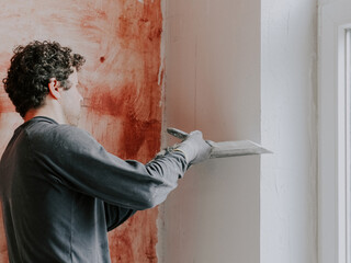A young caucasian guy putty with a large spatula on the wall near the window