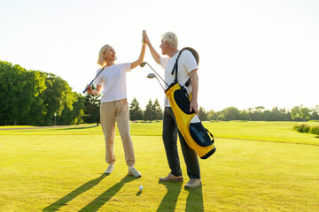 elderly senior couple in uniform celebrating victory and success in golf game and giving high five, old man and woman playing golf on golf course at sunset and doing outdoor sports