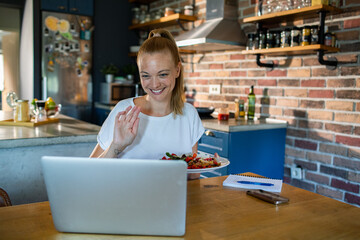 Smiling woman eating salad while video chatting from home kitchen