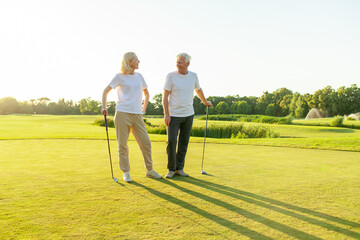 Fototapeta premium old senior couple in white clothes playing golf on golf course and chatting at sunset, elderly man and woman actively relaxing outdoors and laughing, pensioners on vacation