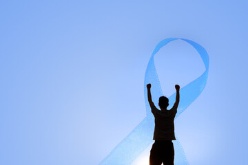 Silhouette of a man raising his arms in front of a large blue ribbon on a clear sky background,...
