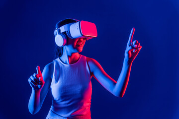 Smart female stand with surrounded by cyberpunk neon light wear VR headset connecting metaverse, futuristic cyberspace community technology. Woman using finger pointing virtual object. Hallucination.