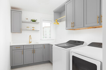 A laundry room with grey cabinets, white washer and dryer appliances, tile flooring, white countertop, and gold faucet and hardware. No brands or labels.