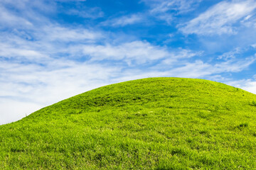 Lush green grassy hill under a clear blue sky with white clouds.
