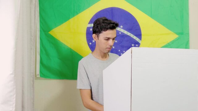 Jovem homem brasileiro na cabine de vota&ccedil;&atilde;o escolhendo o seu candidato na urna eletr&ocirc;nica brasileira. Elei&ccedil;&otilde;es presidenciais e municipais no Brasil. No fundo, a bandeira do Brasil decora o local.