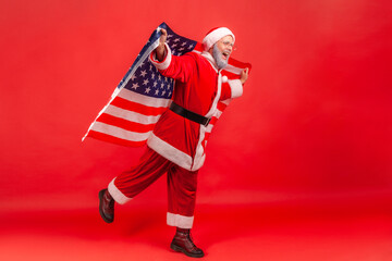 Full length portrait of elderly man with gray beard wearing santa claus costume standing on one leg, pretending to fly with USA flag in hands. Indoor studio shot isolated on red background.