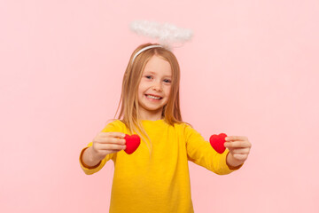 Portrait of smiling delighted blonde little girl with nimb over head showing small hearts looking at camera with happiness, wearing yellow jumper. Indoor studio shot isolated on pink background.