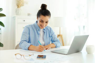 Positive successful motivated arabian or indian woman in a blue shirt, sits a table, working on the laptop, develop new digital project, planning marketing strategy, takes notes in a notebook, smiling