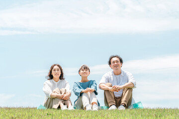 青空の見える公園・丘に座る家族・親子（父親・母親・娘）
