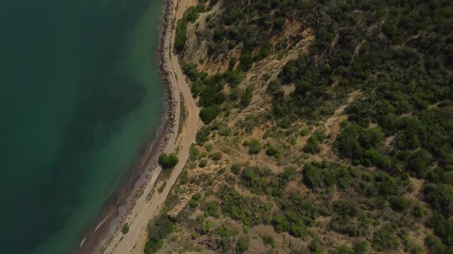 Aerial tracking by drone of a compact blue car, rolling on the ridge of a mountain, with arid vegetation and beach in the background