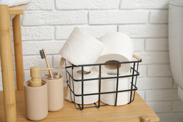 Basket with toilet paper rolls and bath supplies on wooden shelving unit near light brick wall, closeup