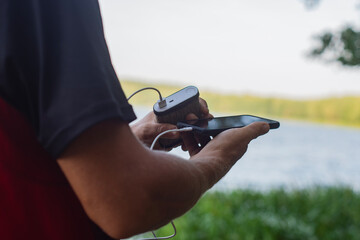 The guy is holding a portable charger with a smartphone in his hand. Man on a background of nature with a greenery and a lake