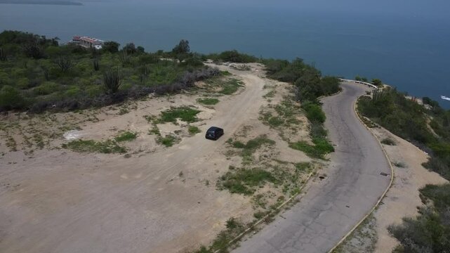 Aerial tracking by drone of a compact blue car, rolling on the ridge of a mountain, with arid vegetation and beach in the background