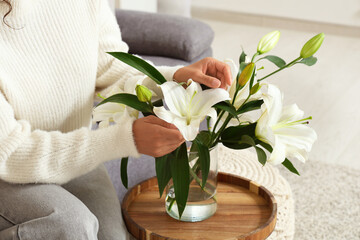 Beautiful young African-American woman with vase of white lily flowers bouquet sitting on sofa at...