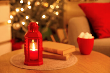 Decoration lantern with books and cup of cacao on table in room decorated for Christmas at night, blurred view