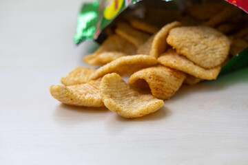 Spicy tomato-flavored potato chips in an open bag. Close-up of tasty snack food on a light wooden surface.