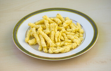 Tasty corn stick chips in a white plate on a wooden background. Kurkure American-style cream and onion flavor chips, a delicious savory snack food.