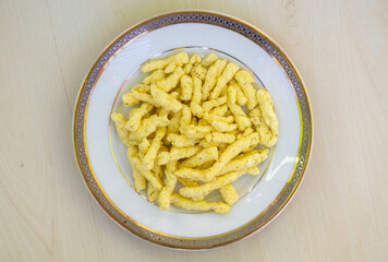 Tasty corn stick chips in a white plate on a wooden background. Kurkure American-style cream and onion flavor chips, a delicious savory snack food. Top view.