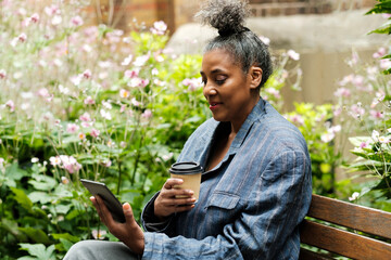 Mature woman on a bench reading an e-book and having coffee.