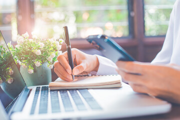 Businesswoman sitting taking notes and searching for information on her smartphone and laptop.