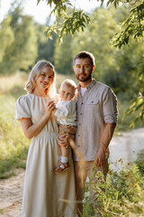 Fototapeta premium A family of three, a man and a woman with a child in their arms, stand in a park in the summer. The concept of a happy family with a small child, care and love.
