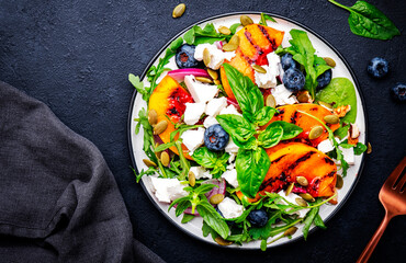 Foodie food salad with grilled peaches, blueberries, red onion, feta cheese, basil and arugula on black background, top view