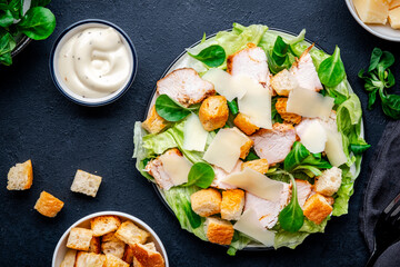 Caesar salad with grilled chicken slices, iceberg lettuce, croutons, parmesan cheese and classic dressing. Black background, top view