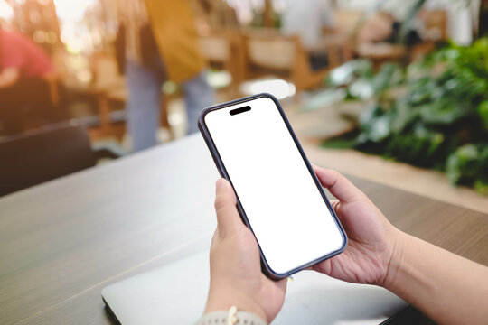 A person holds a smartphone with a blank screen in a casual indoor setting. The background is blurred, showing people and plants, creating a relaxed atmosphere, ideal for browsing or communication.