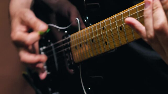 Close-up of a man playing a riff on an electric guitar. Focuses on hands one hand pulling strings with a green plectrum and the other hand pressing them on the fret. Rhythm guitarist