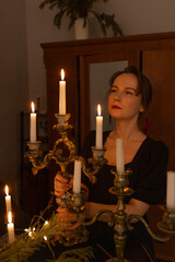 Beautiful young woman extinguishes a candles in candlesticks in dark room. Portrait of girl in black dress in darkness. Fire flames at night, vertical shot