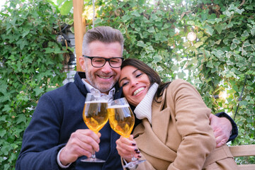 Older couple sharing a romantic evening at a fall outdoor bar, toasting with beer, amidst ivy and lights, exuding joy and affection