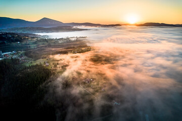Fototapeta premium Stunning aerial view of sunrise over mountainous landscape, with thick fog blanketing valleys and hills. Sun's golden rays pierce through mist, casting serene glow over the entire scene.