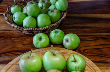 Green apples on a wooden cutting board sitting on a wooden table and some in a basket
