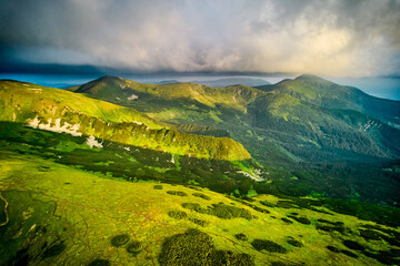 Obraz premium Breathtaking aerial view of green, rugged mountain peaks under dramatic sky. Scene captures contrast between vibrant landscape and moody clouds, with sunlight breaking through. Carpathians, Chornogora