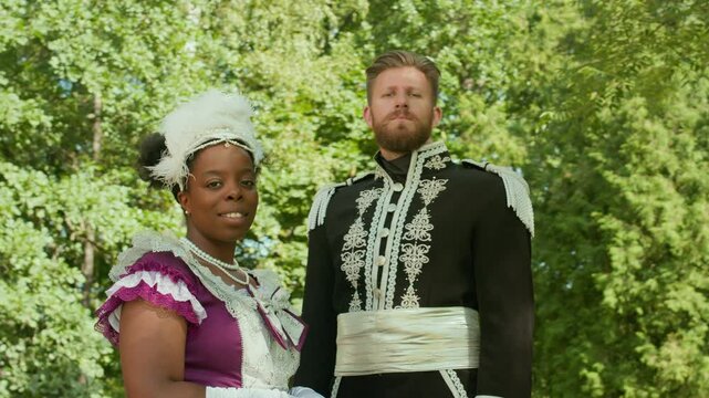 Portrait of Caucasian man in ornate ceremonial uniform with silver embroidery and Black woman in vibrant period dress and feather fascinator posing for camera outdoors against lush tree