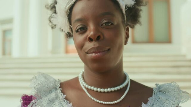 Tilt up portrait shot of Black aristocratic woman wearing period dress, white gloves, pearl necklace and feather fascinator standing outdoors and smiling at camera