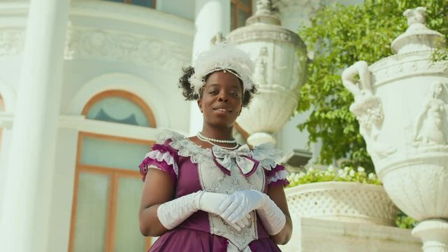 Low angle arc shot of young Black lady wearing feather fascinator, white gloves and vintage dress with lace trim standing in front of historic building and posing for camera with smile