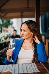 Young beautiful adult woman wearing jacket sitting next to the table in street cafeteria and hold glass of sparkling wine drink romantic date or businesswoman rest