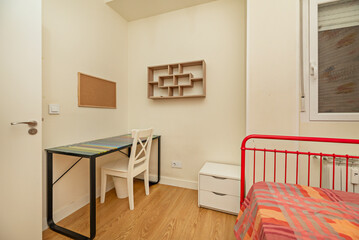 A student bedroom with black metal desk, white wooden chair and red metal bed