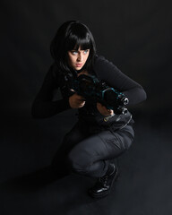 Full length portrait of beautiful black female model wearing black sci fi futuristic dystopian leather bodysuit costume. Kneeling pose, holding gun weapon. isolated dark studio background moody silhou