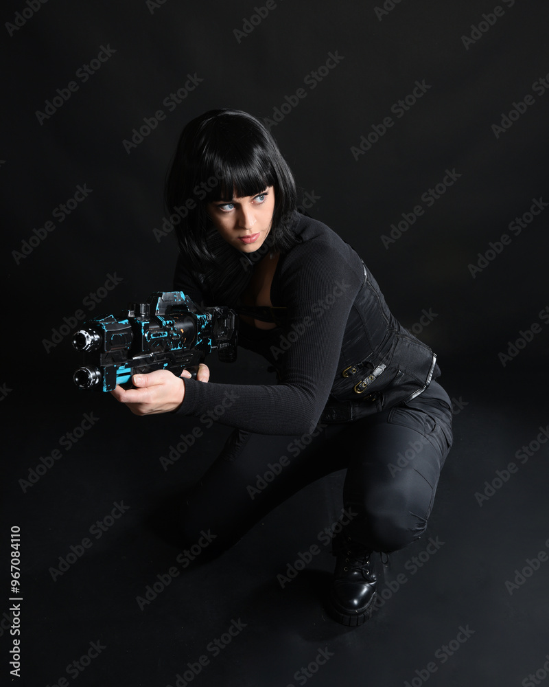 Poster Full length portrait of beautiful black female model wearing black sci fi futuristic dystopian leather bodysuit costume. Kneeling pose, holding gun weapon. isolated dark studio background moody silhou